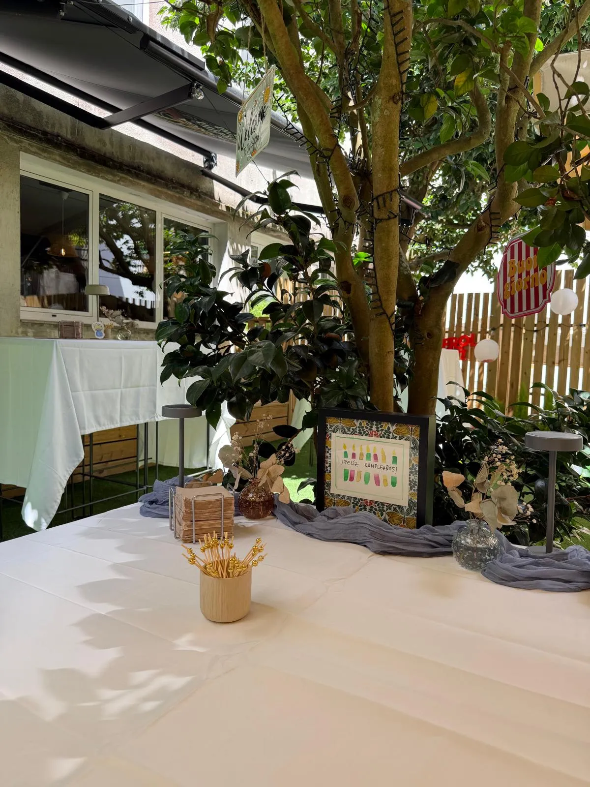 Mesa de cumpleaños montada en la terraza, con cartel de felicitación, camino de mesa gris y eucalipto seco